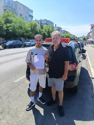 Cursant admis la examenul auto, cu foaia în mână, lângă mașina școlii