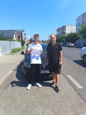 Cursant admis la examenul auto, cu foaia în mână, lângă mașina școlii