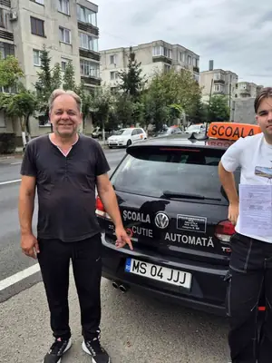 Cursant admis la examenul auto, cu foaia în mână, lângă mașina școlii